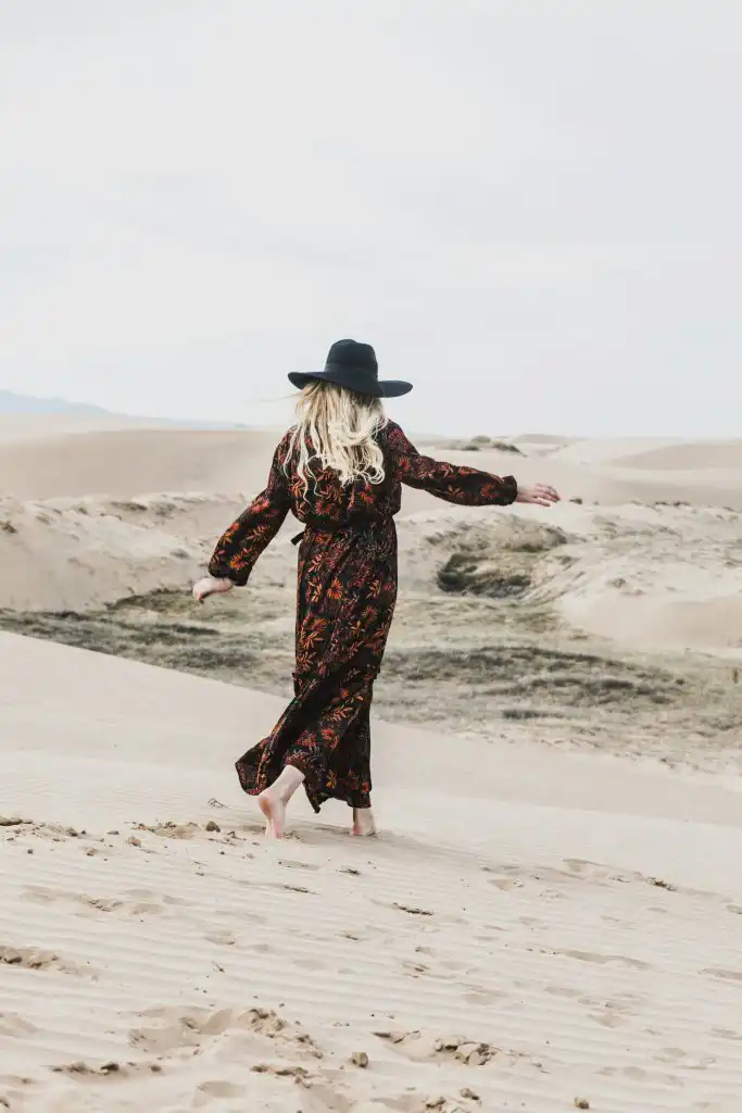Woman in a long dress and hat walking in sandy desert landscape.